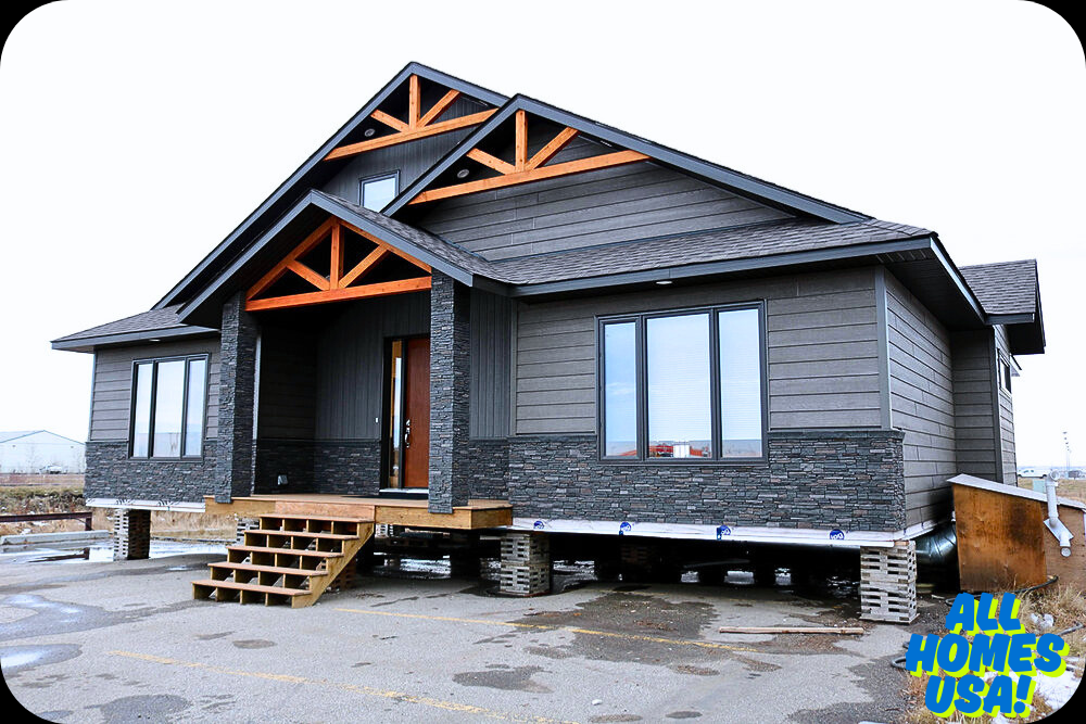A modern modular home on raised supports, featuring dark siding, stone accents, and wooden trim, with temporary steps leading to the front door. "All Homes USA!" logo in bottom right corner. Image for illustrative purpose.