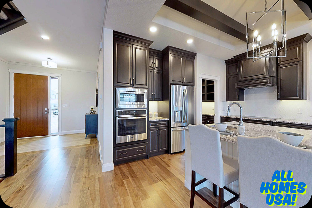Modern kitchen with dark wood cabinets, stainless steel appliances, granite countertops, and white bar stools. Entrance visible with wooden floor and brown door. Bright and welcoming ambiance. Image for illustrative purpose.