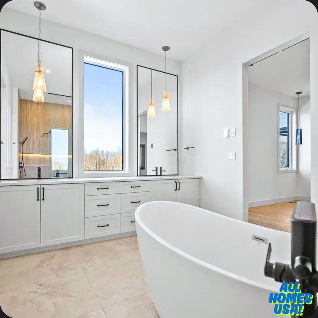 Modern bathroom with a freestanding white tub, dual sinks on a gray vanity, black fixtures, and pendant lights. A large window shows a serene outdoor view. Image for illustrative purpose.