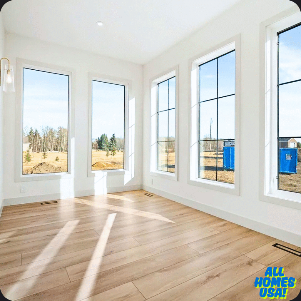Bright room with large windows, white walls, and light wood flooring. Sunlight streams in, casting shadows, with a tranquil, airy feel. Logo reads "All Homes USA!” Image for illustrative purpose.