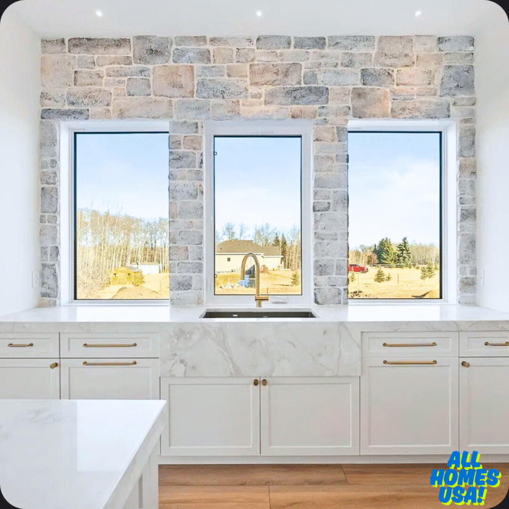 Modern kitchen with white cabinets and marble countertops. A stone wall features three large windows showcasing a scenic outdoor view. Bright and inviting. Image for illustrative purpose.