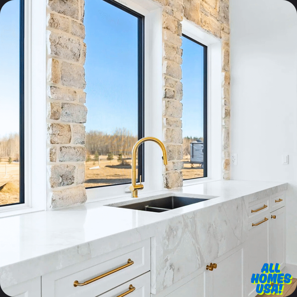 Modern kitchen with white marble countertops, a stainless steel sink, and a gold faucet. Large windows with stone trim show a sunny outdoor view. Image for illustrative purpose.