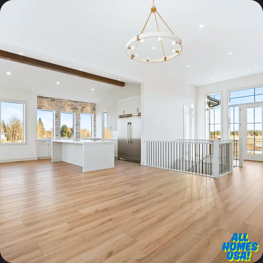 Spacious, sunlit kitchen with wooden floors, large windows, white island, stone accents, stainless steel fridge, modern chandelier, and open layout. Image for illustrative purpose.