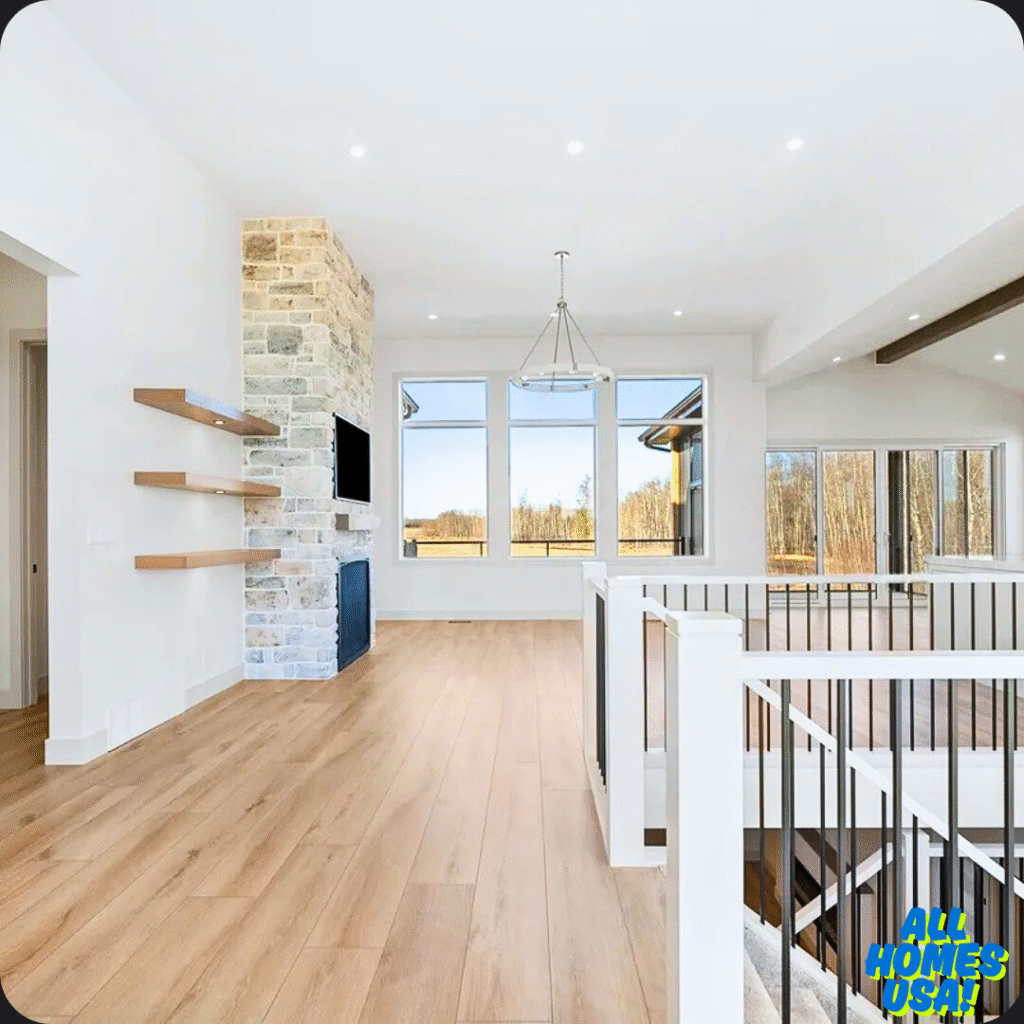 Spacious living room with light wood floors, stone fireplace, and floating shelves. Large windows offer scenic views; sliding doors lead outside. Image for illustrative purpose.