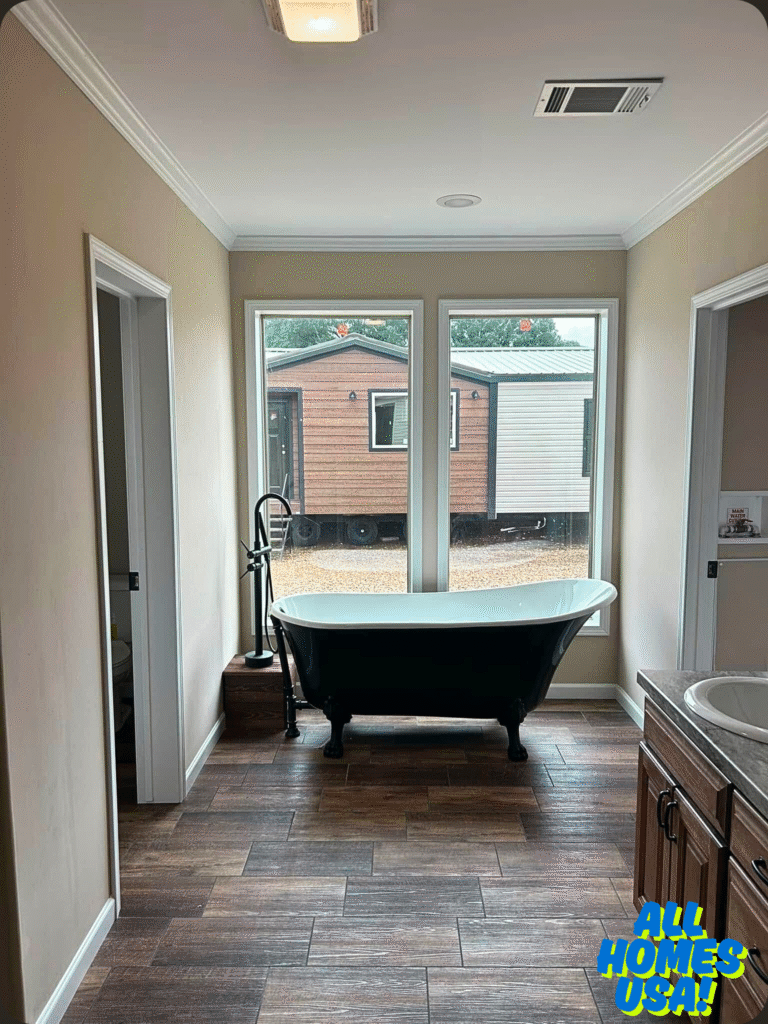 Bathroom features a vintage clawfoot bathtub by large windows, wood flooring, and a brown vanity. Outside, a brown mobile home is visible. Cozy ambiance. Image for illustrative purpose.