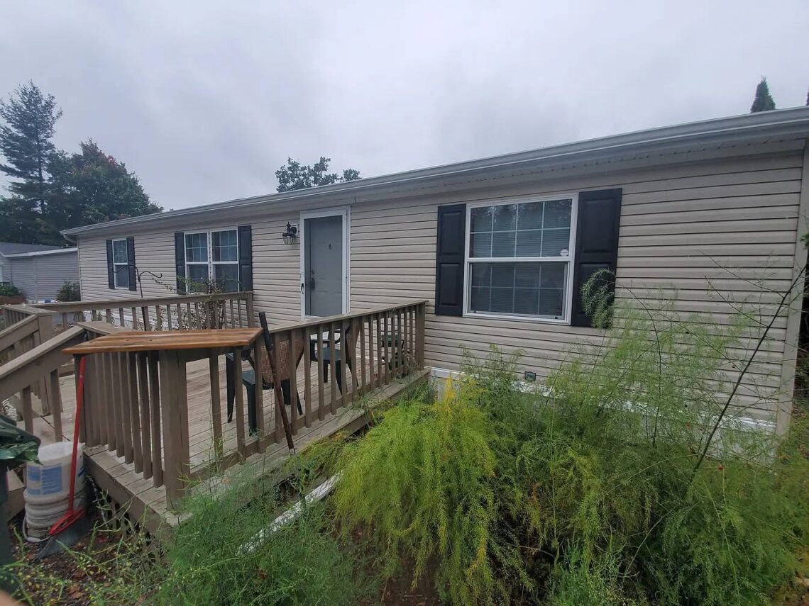 Mobile home with wooden deck, greenery, and overcast sky.