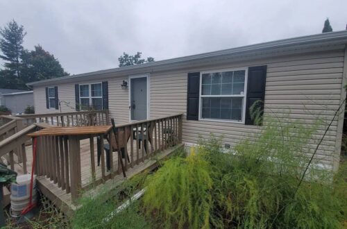 Mobile home with wooden deck, greenery, and overcast sky.