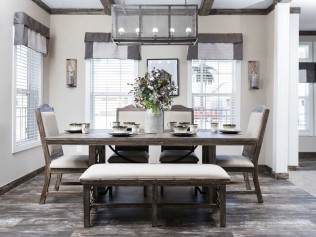 A cozy dining room with a wooden table set for six. Beige chairs and bench surround the table, a vase with flowers at center. Light streams through large windows.