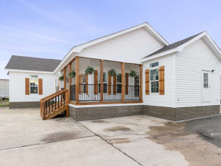 Small white house with a covered wooden porch adorned with hanging plants. The structure has brown shutters and a small ramp leading to the front door. Quiet, suburban setting.