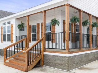 A white house with a wraparound porch featuring wooden columns and railings. Four hanging plants add greenery. Stairs lead to the porch, giving a welcoming feel.