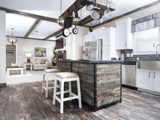 Rustic kitchen with wooden beams, farmhouse sink, and central island with stools. Hanging pots above. Bright, cozy living area visible in background.