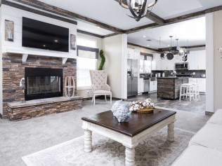 Spacious living room with stone fireplace, wall-mounted TV, and elegant decor. Open layout leads to a modern kitchen. Bright, welcoming atmosphere.