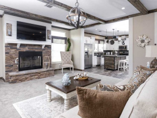 A cozy living room features a stone fireplace, wall-mounted TV, and rustic decor. An adjoining kitchen with modern appliances is visible, fostering warmth and charm.