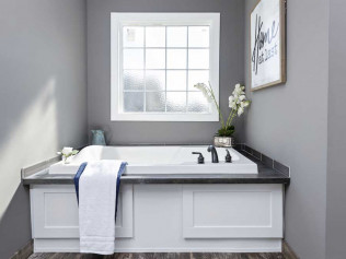 A modern bathroom with a white bathtub next to a large window. Gray walls, wooden floor, towel, and decorative orchid create a calming, spa-like atmosphere.