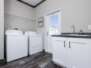 Modern laundry room with a washing machine and dryer side by side on a wood floor. A white cabinet with a sink is on the right, and a door with a window lets in natural light.