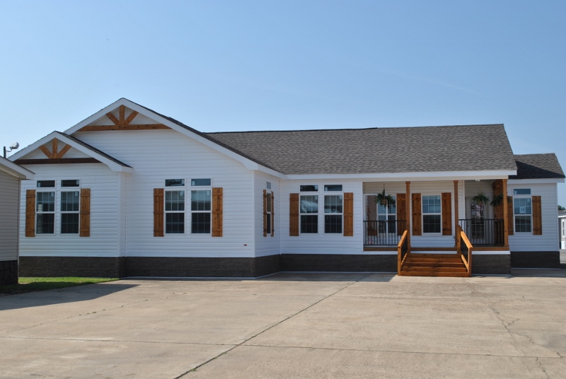 A modern, single-story house with white siding and brown wooden shutters. It features a gabled roof, a small front porch with steps, and hanging plants. The setting is bright and sunny.