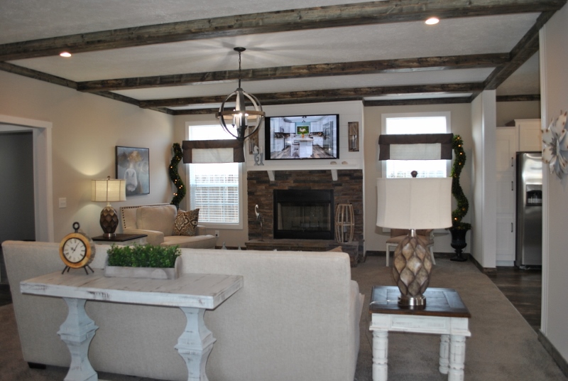 Cozy living room with beige sofas, a stone fireplace, and a TV above. Rustic wooden ceiling beams and a modern chandelier add warmth and style.