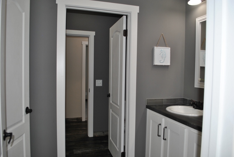 Modern bathroom with gray walls, white trim, and a sink with white cabinets. An open door reveals a view into a hallway with dark wood flooring.