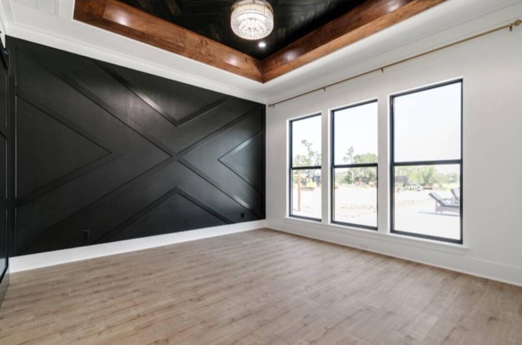 Spacious modern room with light wood flooring, a dark feature wall with geometric patterns, and a tray ceiling with a chandelier. Large windows provide ample natural light. Image for illustrative purposes.