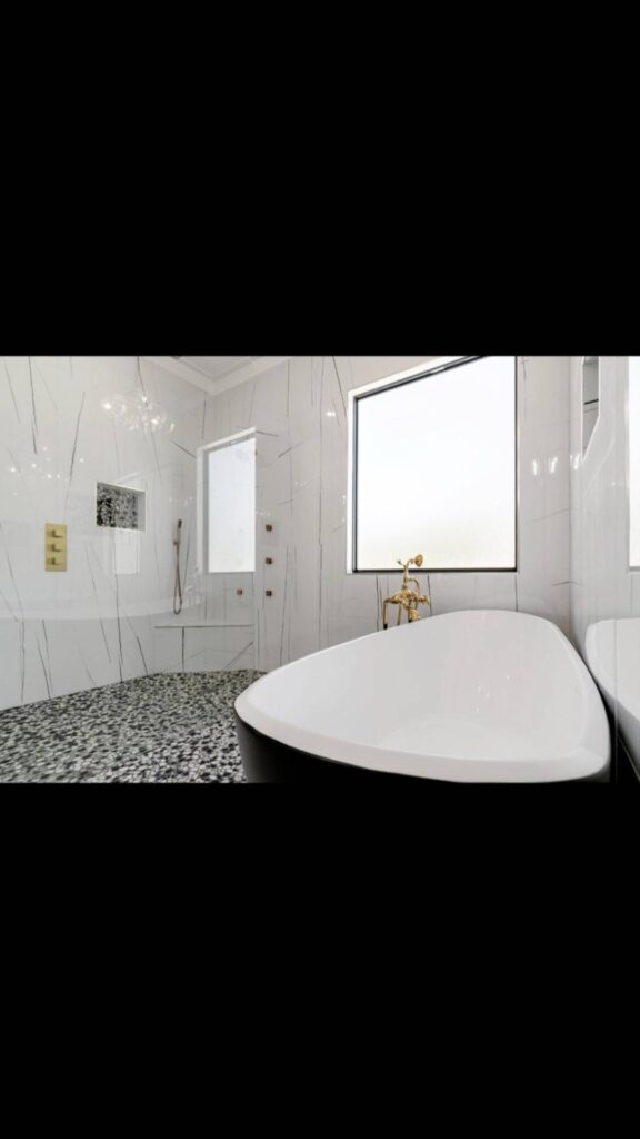 Modern bathroom with a sleek black and white freestanding bathtub beside a large frosted window. Pebble-textured floor, marble walls, and gold fixtures. Luxurious and serene ambiance.