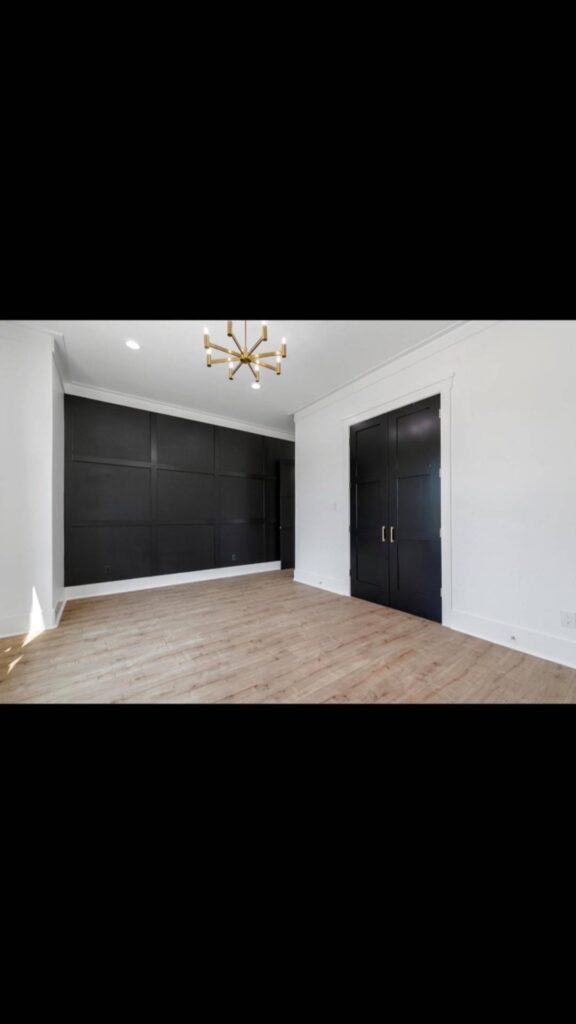 Minimalist room with light wood floors, white walls, and a black accent wall featuring seamless built-in storage. A modern brass chandelier hangs from the ceiling.