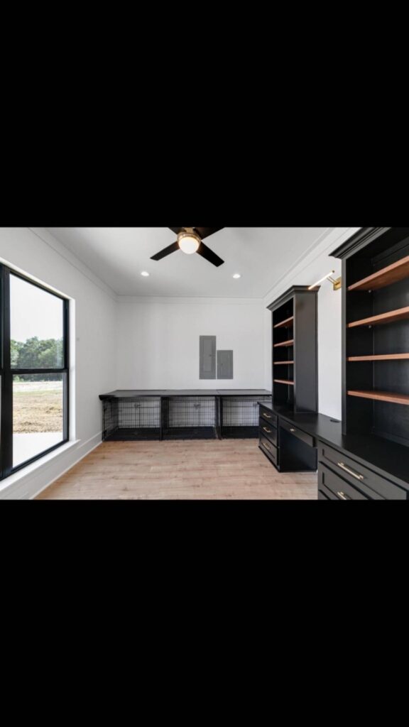 Modern room with black shelving, wooden floors, and a large window offering natural light. A ceiling fan and built-in desks add a sleek touch.