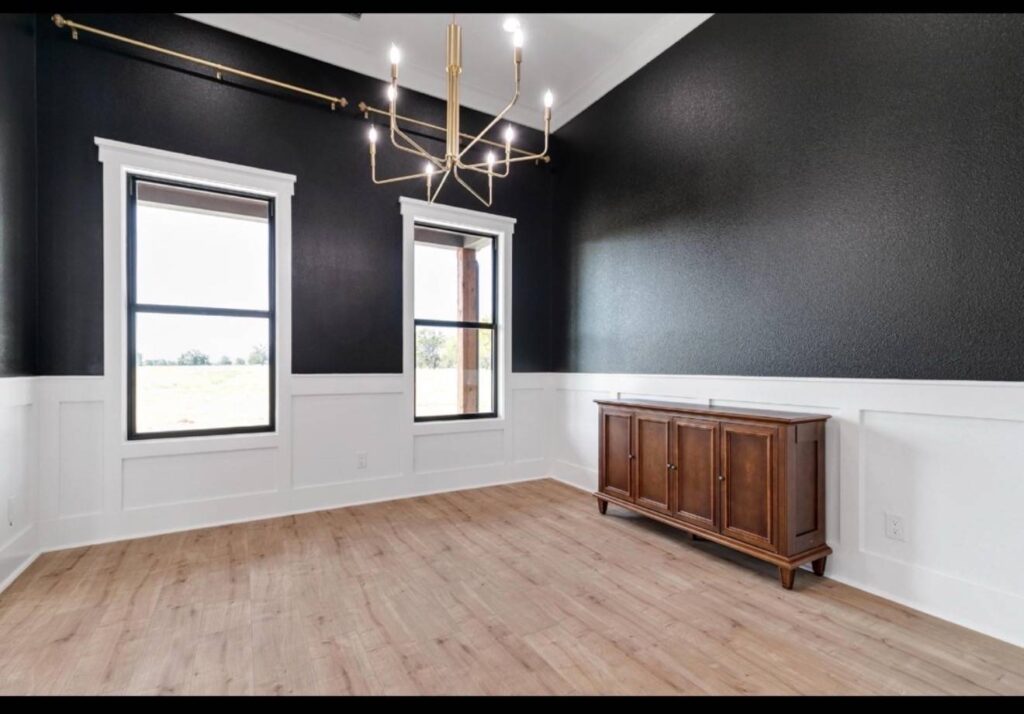 Spacious room with black walls and white wainscoting, featuring two large windows, a modern gold chandelier, wooden sideboard, and light wood floor. Image for illustrative purposes.