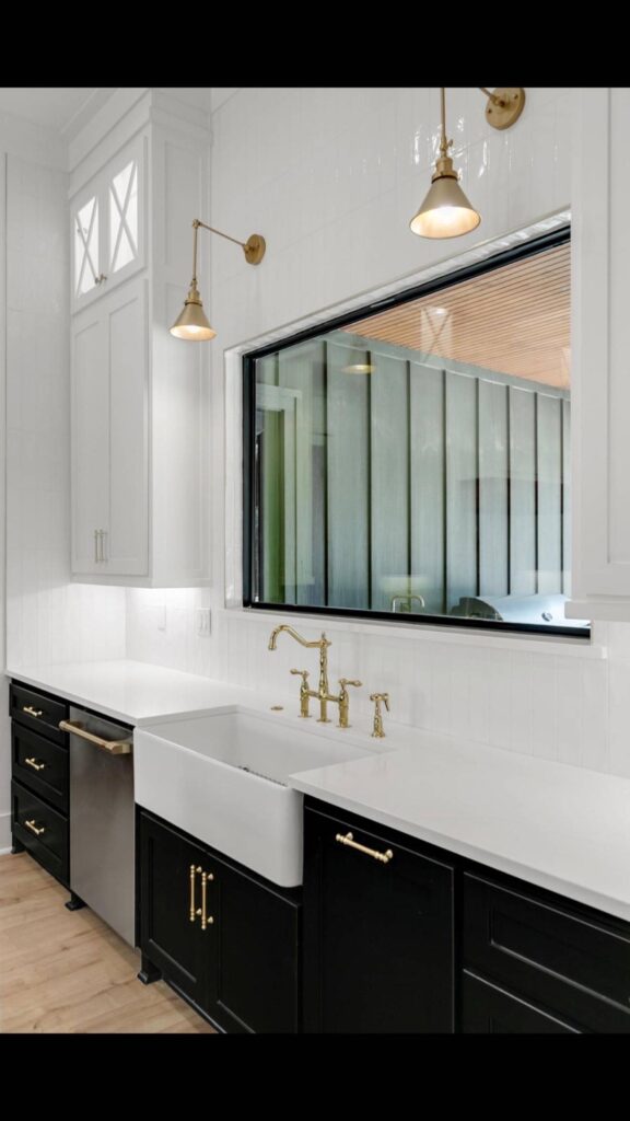 Modern kitchen with black lower cabinets, gold hardware, and a farmhouse sink. Gold faucet, white backsplash, and large window enhance brightness. Image for illustrative purposes.