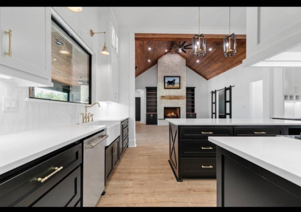 Spacious kitchen with black and white cabinetry, brass fixtures, and pendant lights. Open to a living area with a wooden ceiling and stone fireplace. Image for illustrative purposes.