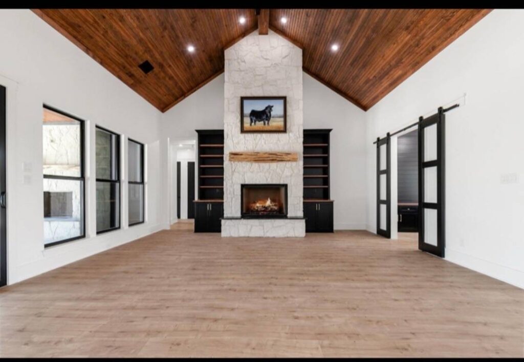 Spacious room with a high, wood-paneled ceiling, light wood floors, and a central stone fireplace. Shelves flank the fireplace, creating a cozy ambiance. Image for illustrative purposes.