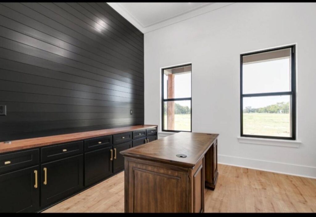 Minimalist office with light wood floor, dark wood desk, and black paneled wall featuring long cabinets. Two windows with greenery view enhance brightness. Image for illustrative purposes.