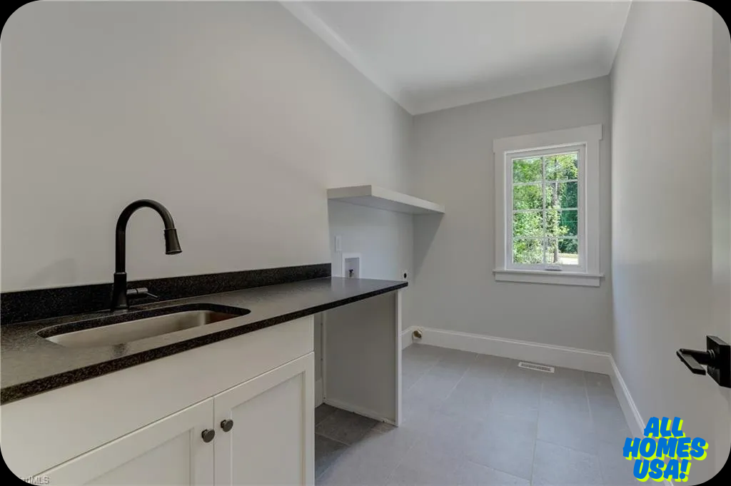 A bright utility room features light gray walls, a sleek black countertop with a sink, white cabinetry, and a window revealing lush greenery outside. Image for illustrative purposes.
