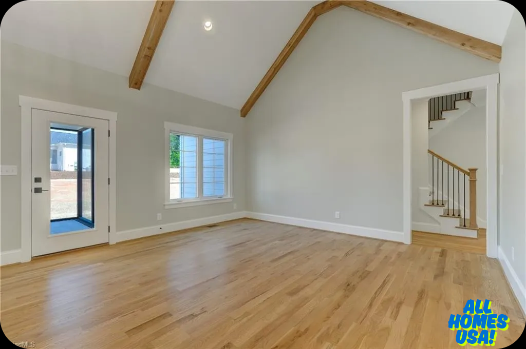 Spacious, empty room with light wood floor, vaulted ceiling with exposed beams, white walls, and two large windows. An open staircase is visible. Image for illustrative purposes.