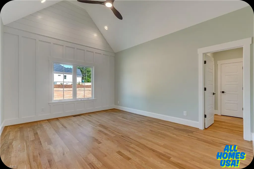 Spacious, empty room with light wood flooring, vaulted ceiling, and soft green walls. Large window and modern ceiling fan convey airy, serene feel. Image for illustrative purposes.