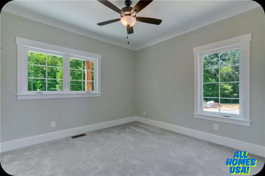 Empty room with gray walls and carpet, featuring a ceiling fan and two windows showing green trees outside. Bright and airy atmosphere. Image for illustrative purposes.