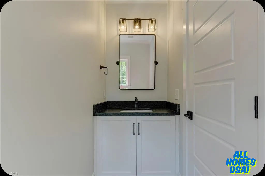 Small, well-lit bathroom with a sleek black granite countertop, white vanity, modern rectangular mirror, and stylish three-bulb light fixture. Image for illustrative purposes.