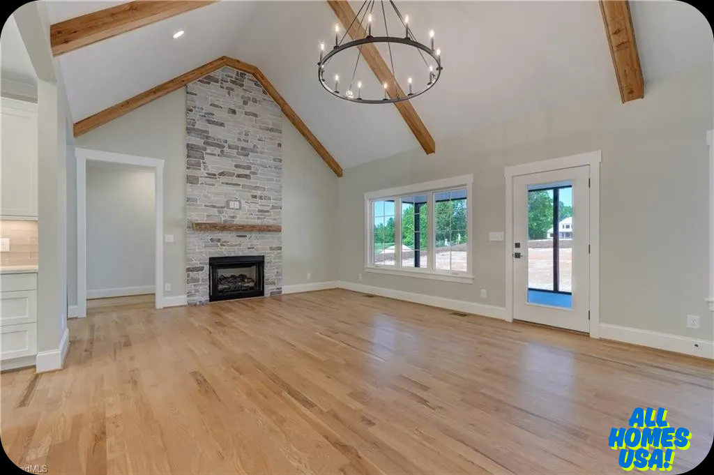Spacious living room with vaulted ceiling, rustic wooden beams, stone fireplace, and sleek chandelier. Large windows and door offer natural light. Image for illustrative purposes.
