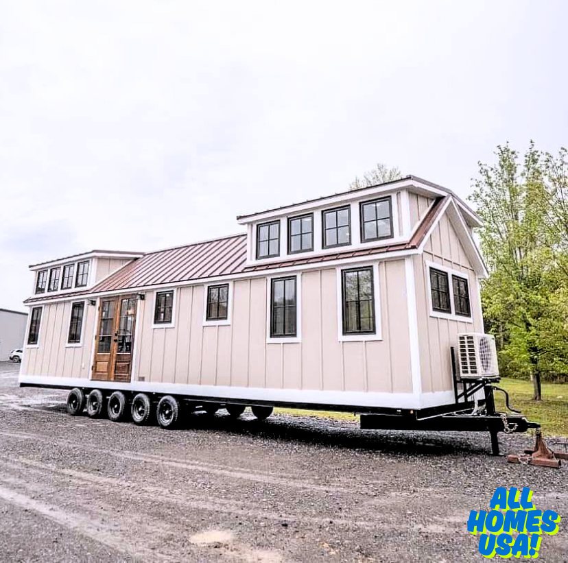 A beige tiny house with black-framed windows sits on a multi-axle trailer. It features a wooden door and a sloped roof, exuding a cozy, modern vibe. Image for illustrative purposes.