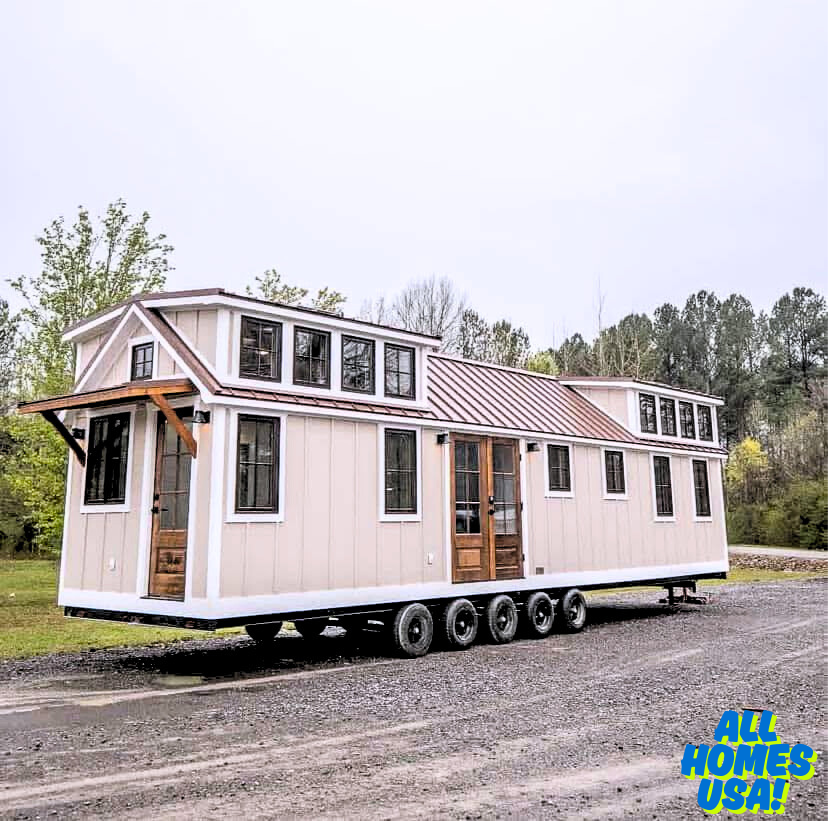 A beige tiny house on wheels with multiple windows and a red metal roof sits on a gravel path, surrounded by greenery and trees under an overcast sky. Image for illustrative purposes.