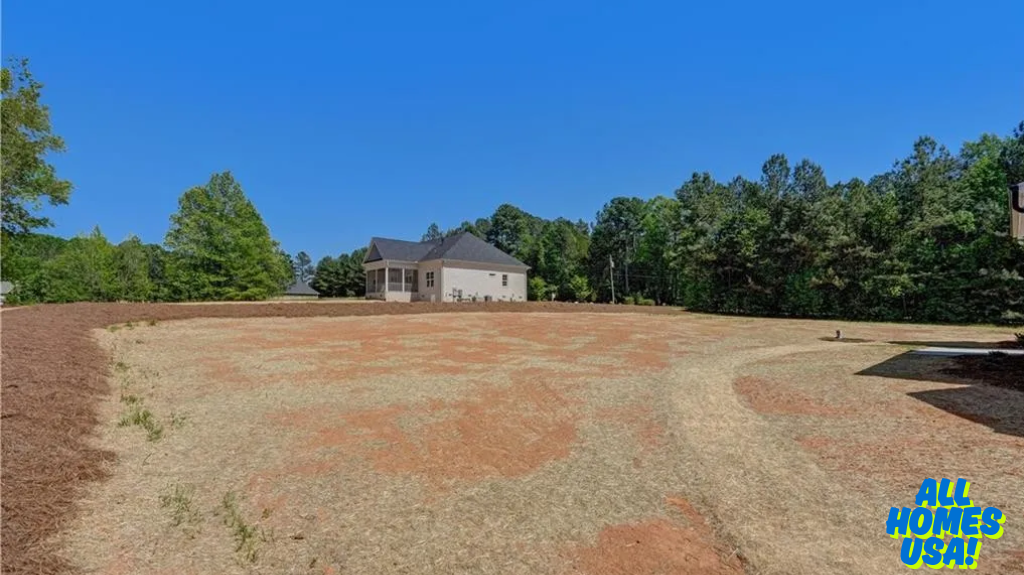 Expansive, empty field with dry grass and sparse vegetation, bordered by lush green trees. A single house with a gray roof is visible in the distance under a clear blue sky. Image for illustrative purposes.