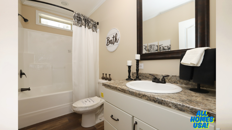 A small, modern bathroom with beige walls features a white bathtub and shower, a granite countertop with a white sink, black fixtures, and a round "Bath" sign on the wall. A white curtain with a floral pattern, candles, and a dark wooden mirror add elegance. The overall tone is clean and inviting. Image for illustrative purposes.