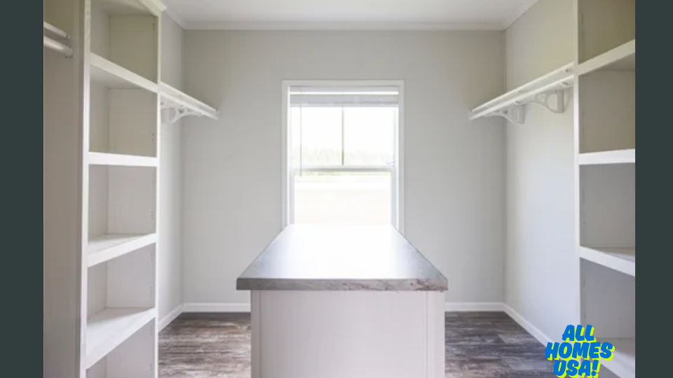 A bright, spacious walk-in closet with light gray walls, featuring white open shelves and hanging rods. A central island with a gray countertop and a window fills the room with natural light, creating an organized and airy feel. Image for illustrative purposes.