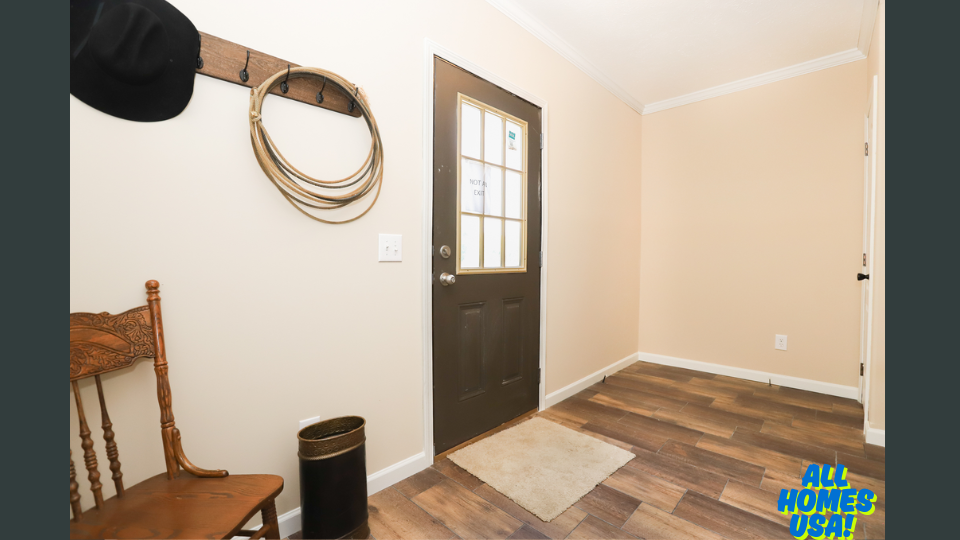 A cozy entryway with a wooden floor and light beige walls. A chair and a wall-mounted rack with hats and ropes add rustic charm. A dark door with glass panels and a small mat complete the scene. Image for illustrative purposes.