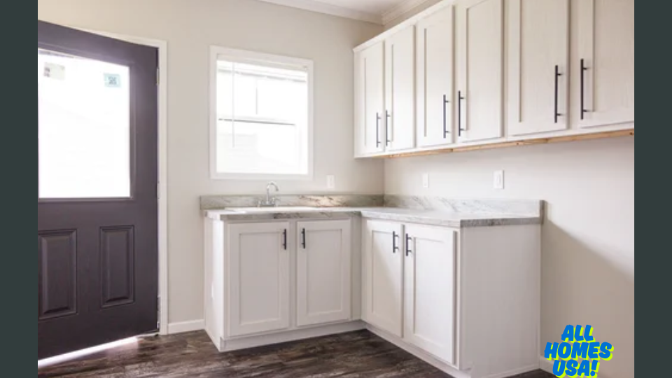 Bright kitchen with white cabinets, marble countertops, and a single window. Dark door and wood flooring add contrast. Logo "All Homes USA!" in corner. Image for illustrative purposes.