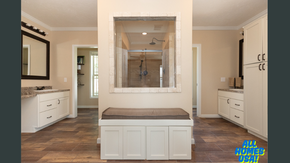 Spacious bathroom with beige walls and wooden tile flooring. Features a central glass shower, white cabinets, large mirrors, and a cushioned bench. Image for illustrative purposes.