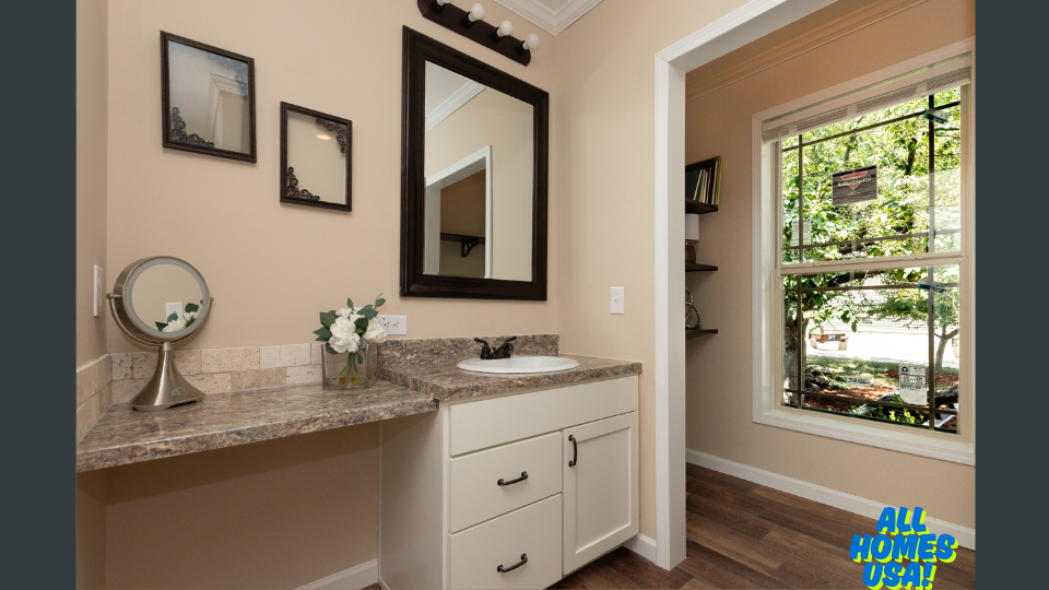 A bathroom with a light beige wall features a granite countertop vanity with a white basin, a round mirror, a vase with white flowers, and two framed mirrors. A large window on the right offers a view of a lush garden. The atmosphere is serene and bright. Image for illustrative purposes.