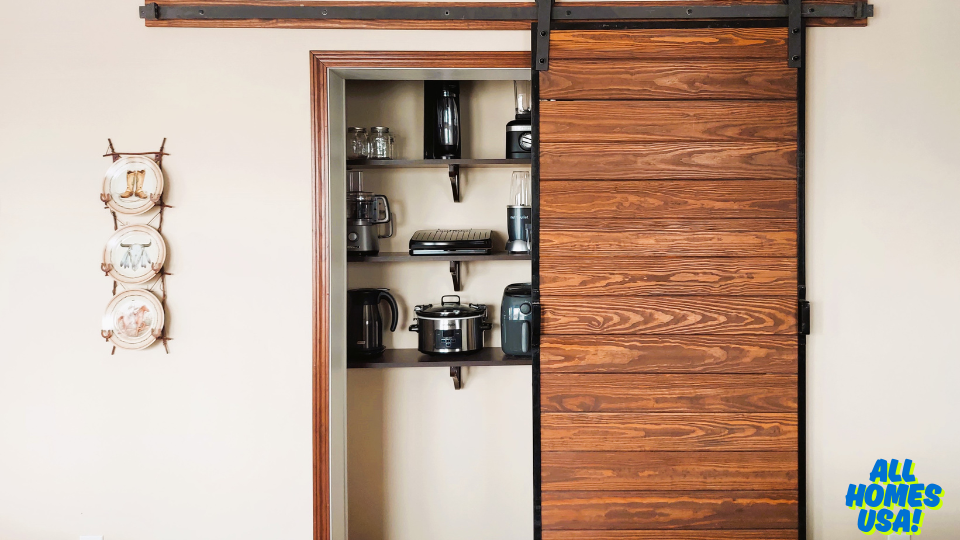 A cozy kitchen features a wooden sliding barn door partially open, revealing shelves with appliances. Four decorative plates adorn the beige wall. Image for illustrative purposes.