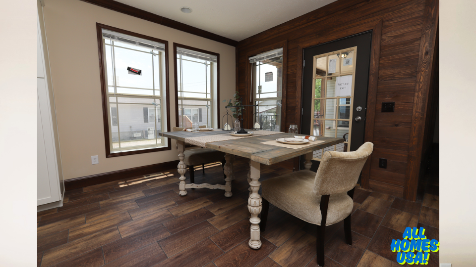 Cozy dining area with rustic wooden table set against a windowed corner. Soft light creates a warm ambiance; chairs are plush, and the flooring is brown wood. Image for illustrative purposes.