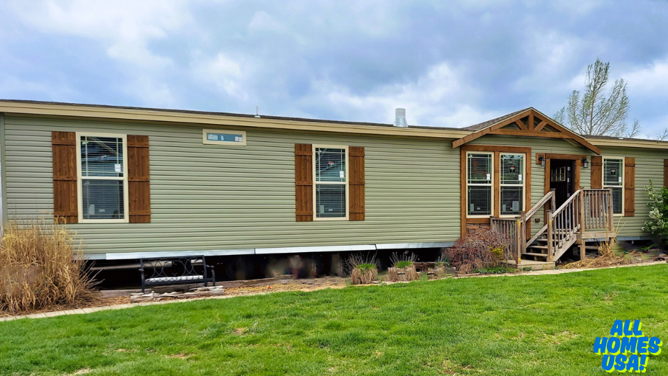 A beige manufactured home with wooden shutters and a porch sits on a green lawn under a cloudy sky. A small bench is visible near the entrance. Image for illustrative purposes.